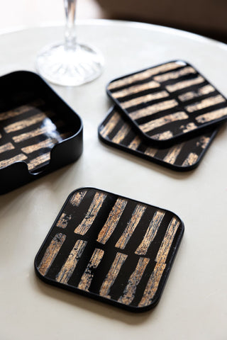 The Set of 4 Black & Gold Brushstroke Coasters With Holder styled on a round table with a glass.