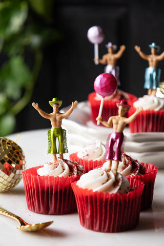 The Set of 4 Cowboy Cake Toppers styled in cupcakes in red cases, on a table with other party accessories.