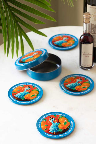 The Set of 4 Flamenco & Oranges Coasters arranged on a white table along with their storage tin, two bottles and the leaves of a plant.