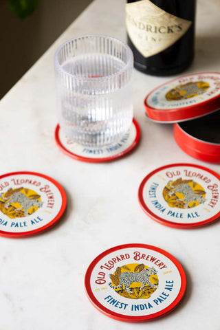 The Set of 4 Old Leopard Brewery Coasters displayed on a white marble table with a glass, bottle of gin and their accompanying storage tin.