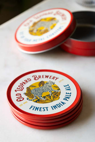 The Set of 4 Old Leopard Brewery Coasters stacked together on a white marble table with their accompanying storage tin in the background with the lid off.