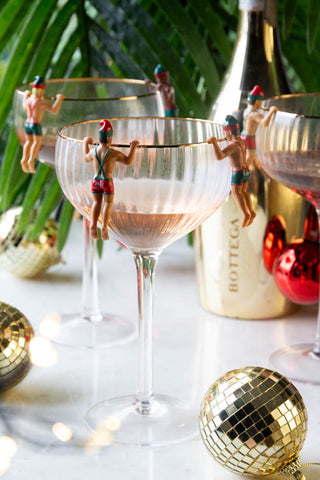 The Set of 6 Christmas Elf Cowboy Drink Markers displayed on the edge of champagne glasses as part of a table decorated with disco balls, a gold bottle and a plant.