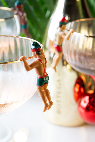 Close-up of one of the Set of 6 Christmas Elf Cowboy Drink Markers displayed on the edge of a champagne glass, with others in the background.