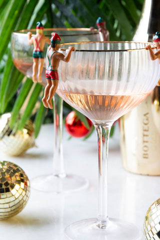 Four of the Set of 6 Christmas Elf Cowboy Drink Markers displayed on the edge of two champagne glasses, styled with a gold bottle, disco ball decorations and a plant.