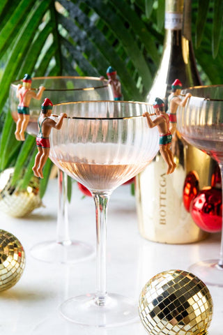 The Set of 6 Christmas Elf Cowboy Drink Markers displayed on the edges of three champagne glasses, styled with a gold bottle, disco balls and a plant.