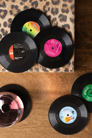 The Set of 6 Vinyl Style Coasters seen from above, styled on a wooden surface with a leopard print book and a glass.