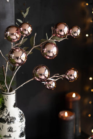 The Shiny Pink Bauble Spray displayed in a vase with some greenery, styled in front of a black wall with some fairy lights and lit pillar candles.