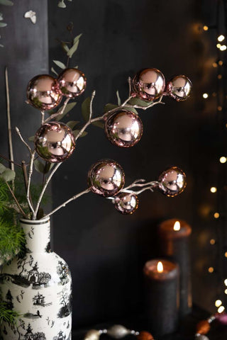 The Shiny Pink Bauble Spray in a vase, styled with greenery, in front of a dark wall with lit candles, fairy lights and a Christmas garland.