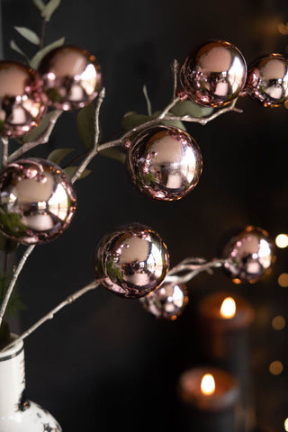 The Shiny Pink Bauble Spray displayed in a vase with some greenery, in front of a dark wall with some lit candles and fairy lights.