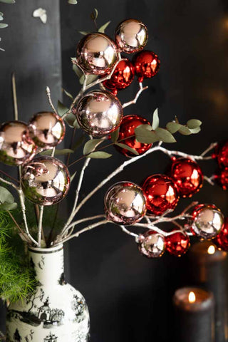 The Shiny Red Bauble Spray styled with the pink version inside a vase with some greenery, in front of a dark wall next to some lit candles.