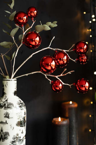 The Shiny Red Bauble Spray displayed in a vase with some greenery, styled in front of a dark wall with lit candles and some fairy lights in the background.