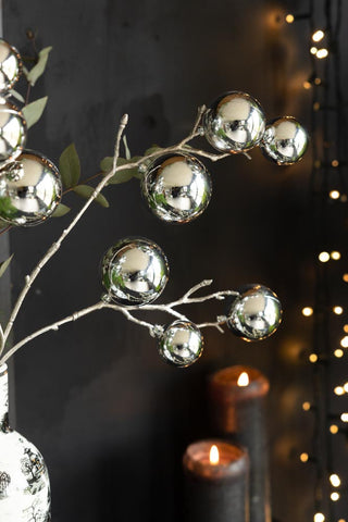 The Shiny Silver Bauble Spray styled with some greenery inside a vase, in front of a black wall with some fairy lights and lit pillar candles.