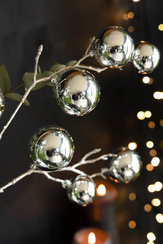 Detail shot of the Shiny Silver Bauble Spray with some greenery, styled in front of a dark wall with some lit pillar candles and fairy lights.