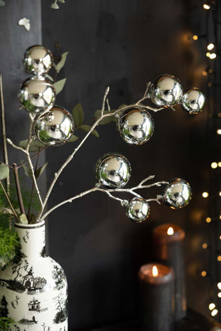 The Shiny Silver Bauble Spray styled in a vase with some greenery, in front of a dark wall with some lit candles and fairy lights.