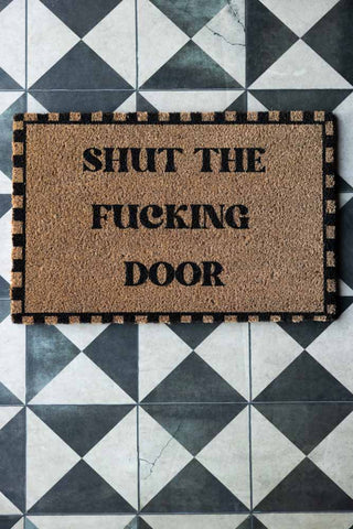 The Shut The Fucking Door Doormat displayed on a geometric floor.