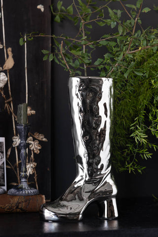 The Fabulous Silver Chrome Boot Vase styled with greenery inside, displayed on a black sideboard with other home accessories.