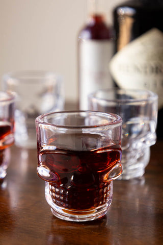 One of the Set of 4 Skull Shot Glasses styled with liquid inside, with the other glasses and some bottles in the background.