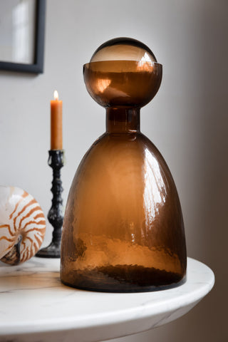 A bronze glass bottle displayed on a white marble table with a shell ornament and a lit candle in a candlestick holder.