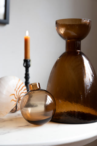 A bronze glass bottle with the round lid removed, displayed on a white table with a shell ornaments and a lit candle in a candlestick holder.