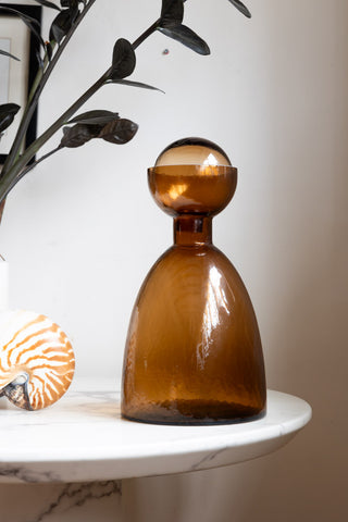 A bronze glass bottle styled on a white marble table with a shell ornament and a plant.