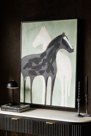 The Stallion Shadow Canvas Art Print seen from an angle, styled on a dark wall above a console table, with a lamp, books and candlestick holders.