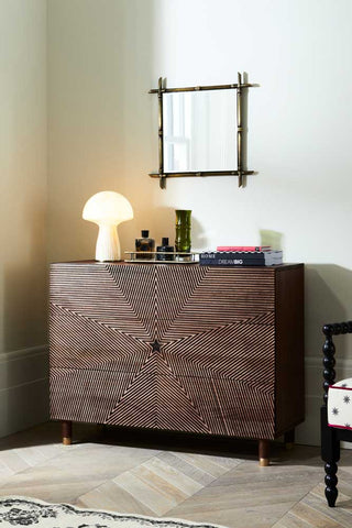The Starburst Chest Of Drawers styled in the corner of a room with various home accessories, a lamp and a chair.
