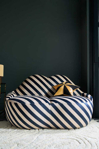 The Stripe Lips Sofa styled in front of a dark wall, with a cushion, side table and lamp.