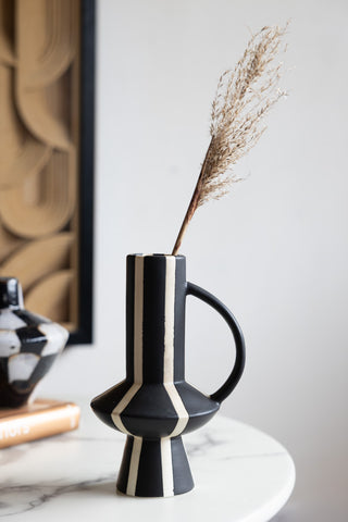 The Tall Striped Black & White Vase With Handle styled with a stem of dried grass inside, displayed on a table.