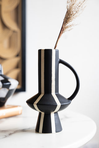 The Tall Striped Black & White Vase With Handle styled with a stem of dried grass inside, displayed on a round table with some other accessories.