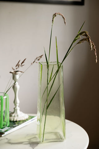 The Tall Textured Glass Green Vase displayed with flowers inside, styled on a table with a vase, candlestick holder and some magazines.