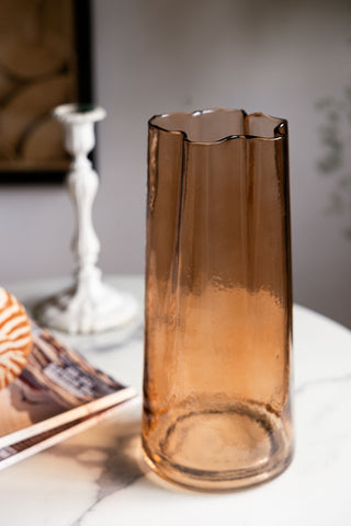 The Tall Textured Sandy Brown Curved Glass Vase displayed on a marble table with magazines and a candlestick holder.