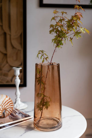 The Tall Textured Sandy Brown Curved Glass Vase displayed with a stem inside, on a table with magazines, a shell ornament and a candlestick holder.