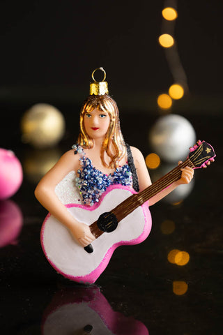 The Taylor Inspired Christmas Tree Decoration styled on a dark surface with baubles and fairy lights in the background.