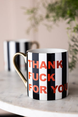 The Thank Fuck For You Stripe Mug on a marble surface, with another mug and a plant in the background.