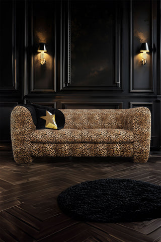 The Bowie Large Sofa In Leopard Love Velvet Natural in front of a dark wall with two lights on, styled with a cushion and a round rug.