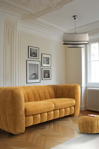 The Bowie Large Sofa In Luxe Needlecord Velvet Golden Glow in a living room in front of a wall with several framed images on, styled with a matching footstool and a pendant light.