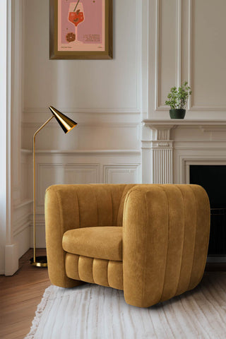 The Bowie Love Seat In Luxe Needlecord Velvet Golden Glow styled in front of a fireplace by a window, with an art print on the wall, plant on the shelf and next to a gold floor lamp.