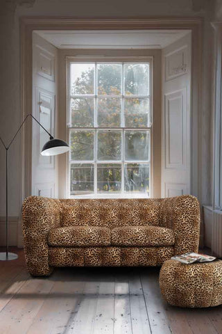 The Bowie Medium Sofa In Leopard Love Velvet Natural styled in front of a large window, with a matching footstool with a magazine on and a large black floor lamp.