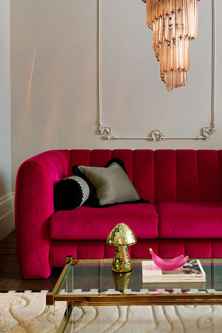 The Bowie Medium Sofa In Luxe Needlecord Velvet Harry's Pink styled in a living room with cushions on, with a large chandelier hanging above and a coffee table with home accessories and magazines on in front.