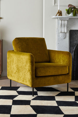 The Edie Needlecord Velvet Green Armchair styled on a black and white rug in a living room, next to a fireplace.