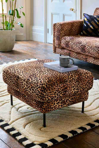 The Grace Foot Stool In Leopard Love Velvet Natural styled in a living room on a rug, with a stack of magazines and a mug on top.