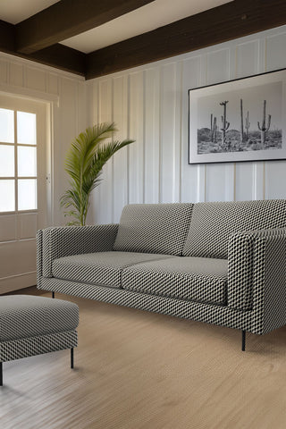 The Grace Large Sofa In Geo Glam Woven Sand & Black styled on a wooden floor in a living room with the matching footstool, in front of a white wall with a cactus art print and a plant.