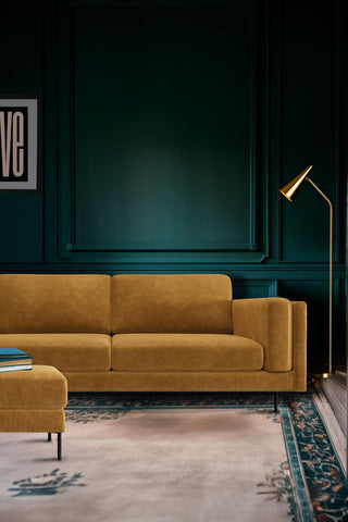 The Grace Large Sofa In Luxe Needlecord Velvet Golden Glow styled in a dark green living room on a decorative rug, with the matching footstool, a floor lamp and an art print.