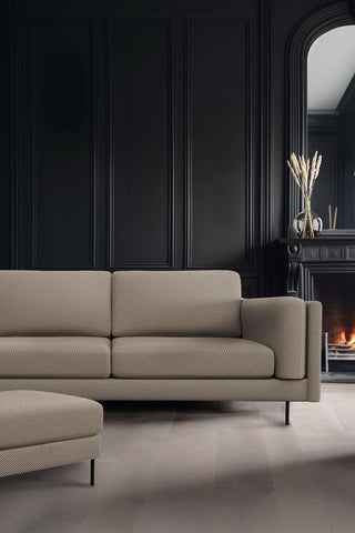 The Grace Large Sofa In Studio Stripe Velvet Sand & Black styled in a living room with black walls, next to the matching footstool, with a lit fireplace and a vase with dried grasses in the background.