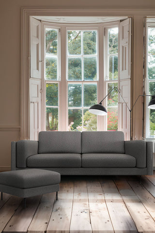 The Grace Large Sofa In Timeless Texture Black Bean styled in front of a large window, with the matching footstool and a floor lamp.