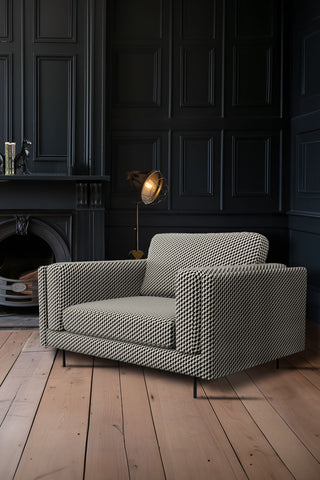 The Grace Love Seat In Geo Glam Woven Sand & Black styled in the corner of a dark room next to a floor lamp and a fireplace.