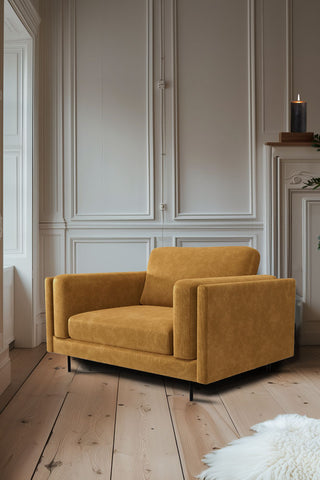 The Grace Love Seat In Luxe Needlecord Velvet Golden Glow styled in the corner of a living room by a window, with a firplace styled with greenery and a lit candle in the background.