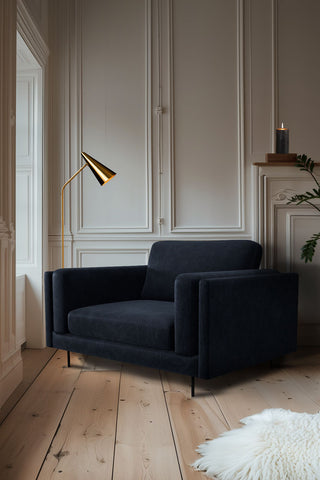 The Grace Love Seat In Luxe Needlecord Velvet Midnight Navy styled in the corner of a room by a window, with a lamp, rug, plant and a lit candle.