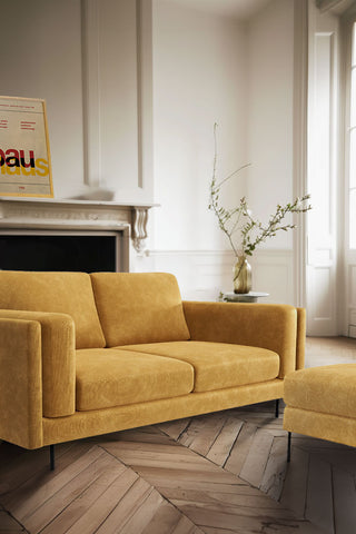 The Grace Medium Sofa In Luxe Needlecord Velvet Golden Glow styled in a living room with the matching footstool, a side table with a vase and an art print on the shelf of the fireplace in the background.