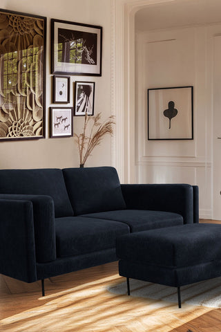 The Grace Medium Sofa In Luxe Kneedlecord Velvet Midnight Navy styled in a living room with the matching footstool, with pampas grass and various art prints in the background.